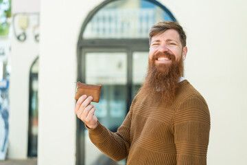 Redhead man with beard holding a wallet at outdoors smiling a lot