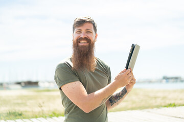 Redhead man with beard holding a tablet at outdoors smiling a lot