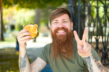 Redhead man with beard holding a burger at outdoors smiling and showing victory sign