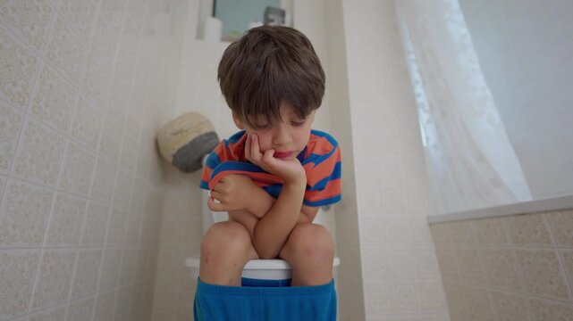 Young boy sitting on toilet in brightly lit bathroom resting chin on hand with bored expression, pants pulled down around ankles