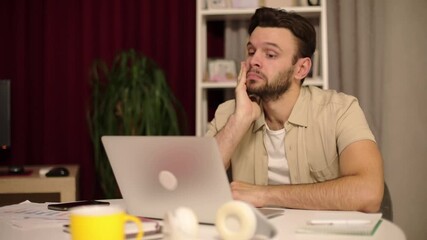 Man sitting at a desk, looking tired and frustrated while working on a laptop. His hand supports his head in exhaustion. Home office scene with warm lighting and scattered objects. - Powered by Adobe