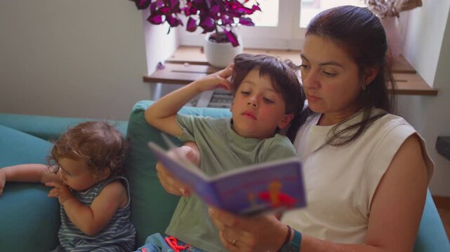 Mother reading storybook to two children on living room couch, toddler distracted while older child leans in, quiet afternoon bonding moment by the window