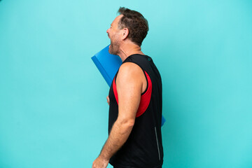 Middle age sport man going to yoga classes while holding a mat isolated on blue background laughing...