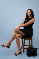 Confident young woman sitting on a stool with legs crossed, wearing a black dress and heels. Studio...