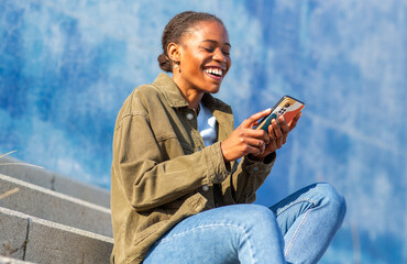 Fototapeta premium African young woman laughing while using smartphone on outdoor steps on blue background