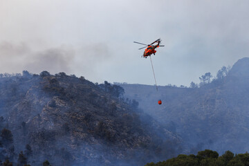 Helicoptero no reconocible con Bambi Bucket se aproxima al fuego en incendio forestal del parque natural de la Font Roja en julio de 2025, Ibi, Espa&ntilde;a