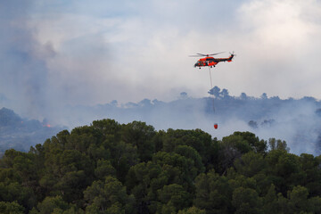 Helicoptero no reconocible con Bambi Bucket se aproxima al fuego en incendio forestal del parque natural de la Font Roja en julio de 2025, Ibi, Espa&ntilde;a