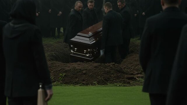 Funeral ceremony showing a wooden coffin being lowered into grave. This funeral ceremony is somber, attended by mourners in black attire, standing in respect and grief.