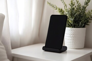 Modern black smartphone standing on its black support on a white bedside table with a decorative plant next to it, in a bright bedroom