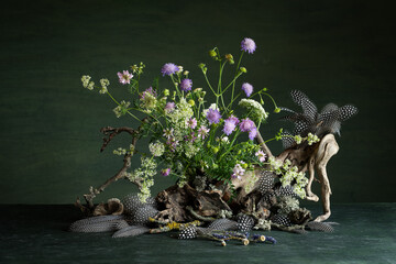 Composition of vibrant wildflowers, delicate guinea fowl feathers, lichens, and tree branches on a dark green background, radiating a mystical and mysterious atmosphere with expressive natural beauty.