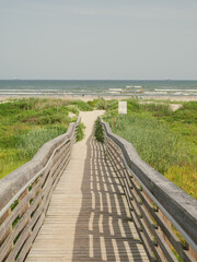 Long wooden boardwalk leads to Galveston beach where people relax, rainbow colorful umbrella. Coastal grass, ocean waves, and sunny skies highlight leisure, nature, Gulf Coast beach activities