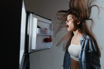 Surprised woman staring into an open refrigerator, her hair blown back, reflecting shock and...
