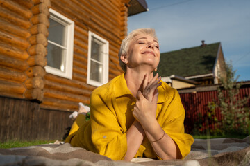 A serene scene of a mature woman dressed in yellow, leisurely relaxing on a blanket in her garden near house, enjoying sun