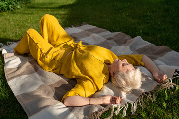 A serene scene of a mature woman dressed in yellow, leisurely relaxing on a blanket in her garden near house, enjoying sun