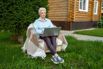 A senior woman is engaged in her work outdoors, using a laptop in a cozy garden environment