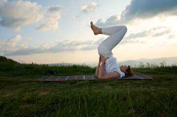 Woman engages in a yoga pose while lying on a mat in a peaceful outdoor location, surrounded by...