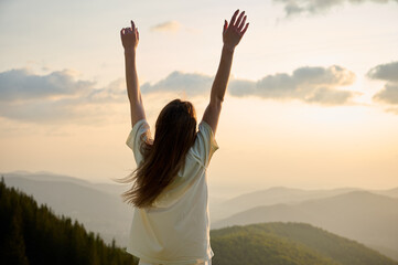A woman stands on a mountain ridge with arms raised high, celebrating the beauty of nature as the sun sets in the background. Surrounding hills are covered in rich greenery, creating serene atmosphere