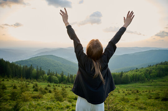 A woman stands on a mountain ridge with arms raised high, celebrating the beauty of nature as the sun sets in the background. Surrounding hills are covered in rich greenery, creating serene atmosphere