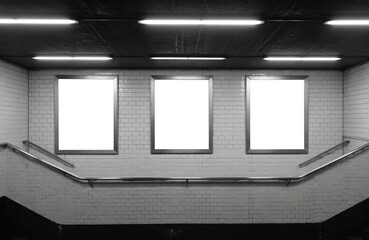 Subway station interior with white wall and three window triptych. Billboards advertising in black and white tunnel. Triangular formation of light reflections. Black metal railing on platform.