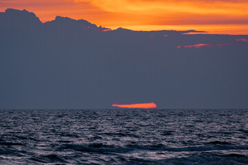 A dramatic sunrise peeks through dark clouds over the sea, casting vivid orange and yellow tones...
