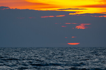 A dramatic sunrise peeks through dark clouds over the sea, casting vivid orange and yellow tones...