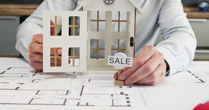 Office worker gently opens tiny front door of house model. Man architect positions little Sale sign neatly on table. Male describes features of house using model