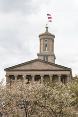 Tennessee State Capitol with flowering cherry blossom trees in the foreground. The American, Tennessee, and General Assembly of Tennessee flags are flying proudly from the top of the capitol.