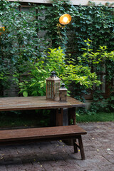  Wooden table and benches with decorative lanterns, surrounded by lush green garden foliage. Soft ambient lighting creates a relaxing outdoor atmosphere perfect for leisure.