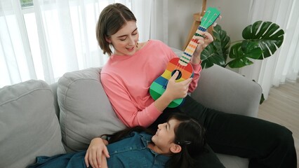Asian cute daughter listening acoustic music while lie on her mother lap at sofa. Caucasian mother...