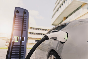 Electric vehicle receiving charge near buildings under sunlight.