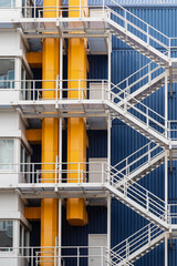Building with a white fire escape along yellow pipes against a blue background in the center of Rotterdam in the Netherlands.