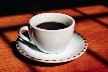 Cup of coffee with a saucer on a wooden table, illuminated by warm sunlight streaming through a window, creating soft shadows in a cozy atmosphere