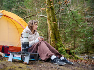 Hiker woman drinking coffee outdoor near the camping tent. A female traveler in the forest in early spring holds hot coffee in a tourist mug.
