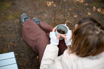 Hiker woman drinking coffee outdoor near the camping tent. A female traveler in the forest in early spring holds hot coffee in a tourist mug.