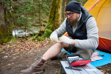 Hiker man with injured leg using first aid kit near the tent in the forest. A stressed trekker complaining alone in the mountain with broken knee.