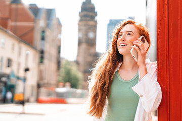Young woman enjoys a pleasant conversation while standing near a red telephone box in an urban setting on a bright sunny day