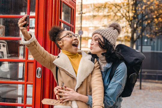 Two friends joyfully take a selfie outside a red telephone booth during a sunny autumn day in an urban setting filled with warm colors and vibrant energy