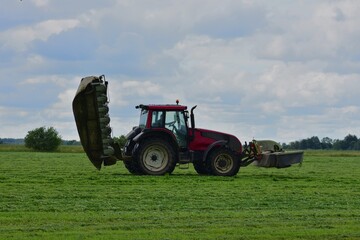Side view of red tractor with raised mowing attachment on a grassy field under a cloudy sky. Farming machinery during seasonal work.