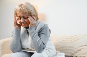 Senior woman sitting on a sofa at home with a headache, feeling pain and with an expression of...