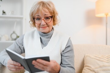 cheerful senior woman sitting on the couch reading a book at home. Education, mature, leisure...