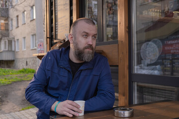 man with a beard sits calmly at an outdoor cafe, holding a cup