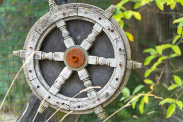 Old Wooden Ship's Wheel. A weathered wooden steering wheel with a rusty center hangs decoratively amidst the greenery. The wheel is reminiscent of times gone by on the high seas.