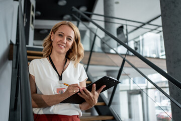 Smiling businesswoman taking notes on clipboard standing on stairs in office building