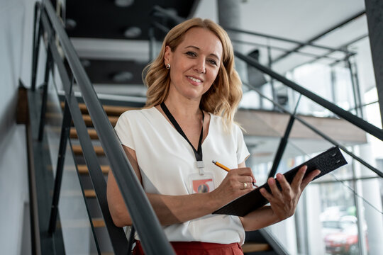 Smiling businesswoman writing notes on clipboard standing on stairs in modern office