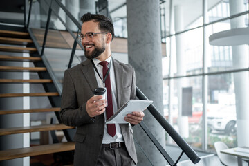 Fototapeta premium Smiling businessman holding tablet and coffee, walking up stairs in office
