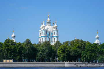 A striking church with blue and white domes rises above lush green trees near a calm river in Saint Petersburg. The clear sky enhances the serene atmosphere of the location