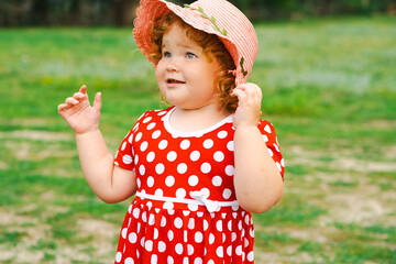 Smiling Child in Red Polka Dot Dress and Pink Hat Enjoys Sunny Day Outdoors in Green Park Setting