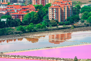 Colorful salt pans reflect apartment buildings and greenery in Cagliari, Sardinia, creating unique blend of urban and natural beauty. Vibrant waters contrast with lush surroundings in Sardinia