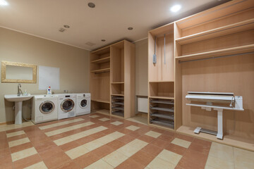 A functional laundry room with two washing machines, a working area, and ample storage space. The room has a warm color palette and is well-lit with bright overhead lights.