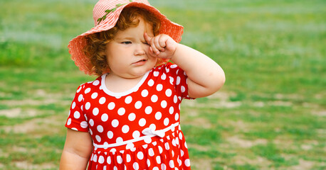 A young child enjoys a sunny day outdoors in a grassy area dressed in a bright red polka dot dress and a charming sun hat playfully touching her face as she explores her surroundings